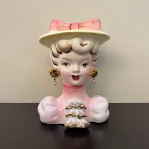 Puede incluir: Jarrón de cerámica vintage con forma de cabeza de mujer. El jarrón está pintado en tonos rosa, blanco y dorado, con un lazo rosa en el sombrero. La mujer tiene pendientes de perlas y lápiz labial rojo. Un artículo decorativo.