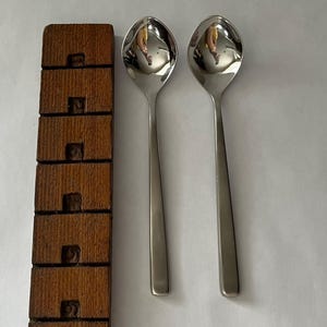 May include: Two shiny, stainless steel spoons with oval bowls and long, slender handles. A wooden spoon rest with carved rectangular cutouts is to the left. The spoons reflect the surrounding environment.