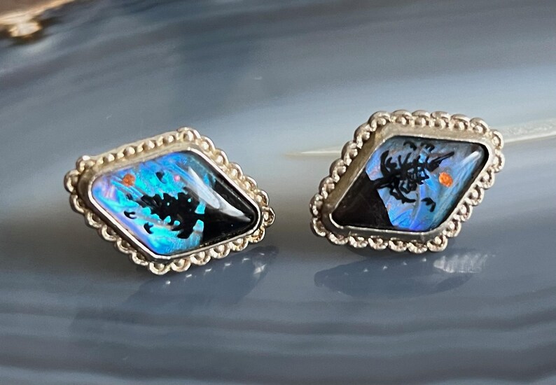 May include: A pair of silver earrings featuring a blue and black butterfly wing design. The wings are set in a diamond-shaped frame with a beaded border.