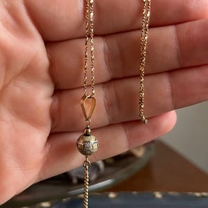 Puede incluir: Un collar de cadena de oro con un colgante de bola texturizada. El colgante tiene una cadena m&aacute;s peque&ntilde;a unida a la parte inferior.