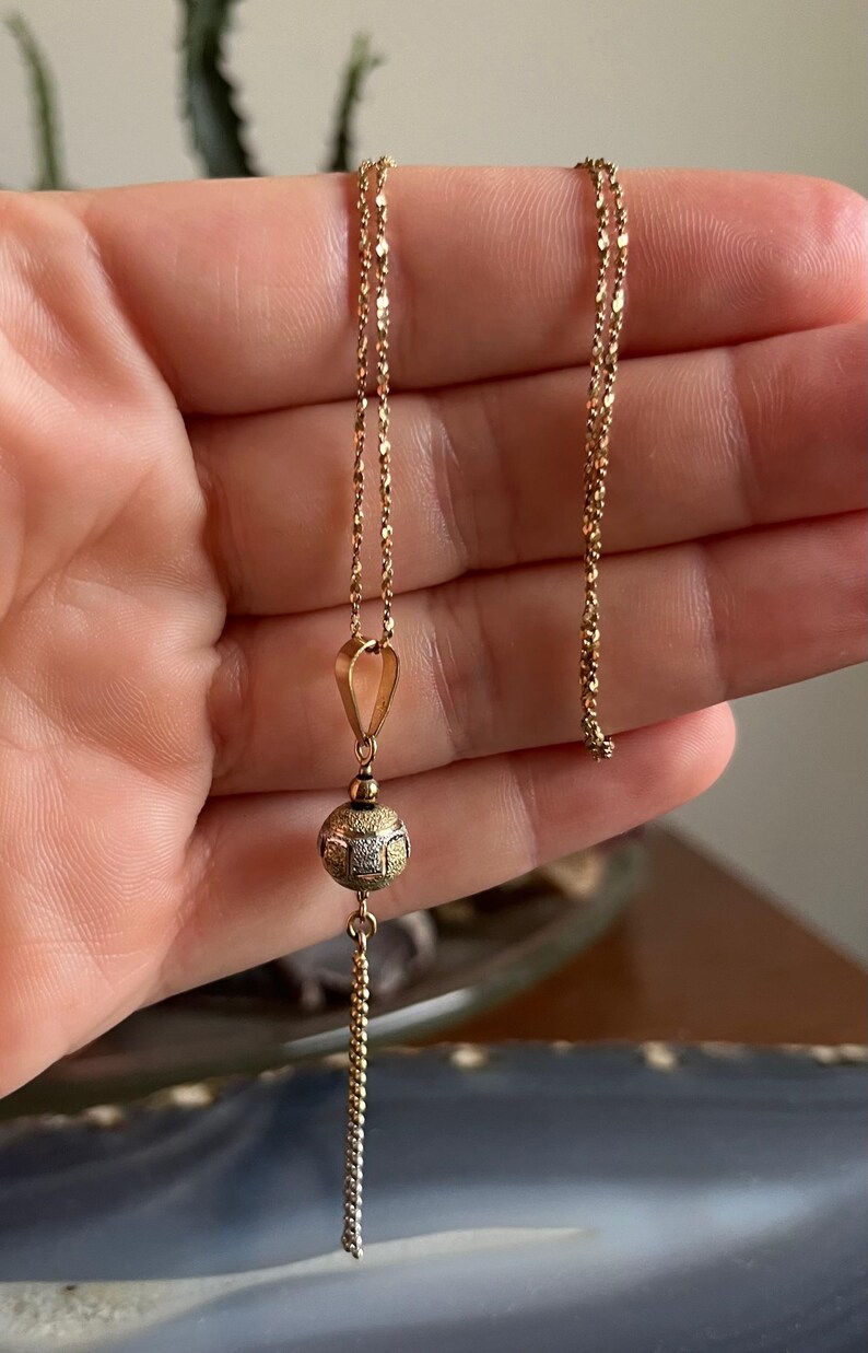 Puede incluir: Un collar de cadena de oro con un colgante de bola texturizada. El colgante tiene una cadena m&aacute;s peque&ntilde;a unida a la parte inferior.