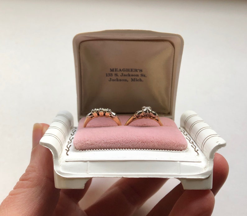 K&ouml;nnte beinhalten: Wei&szlig;e Ringbox in Form einer Couch mit zwei goldenen Ringen mit Diamanten darin. Die Box tr&auml;gt den Text "MEAGHER'S 123 S. Jackson St. Jackson, Mich." auf dem inneren Deckel.