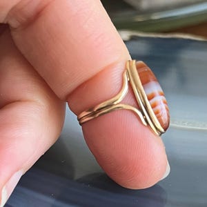 以下が含まれることがあります： 赤と白の瑪瑙の石がベゼルセッティングでセットされた金のリング。リングは指にはめられています。
