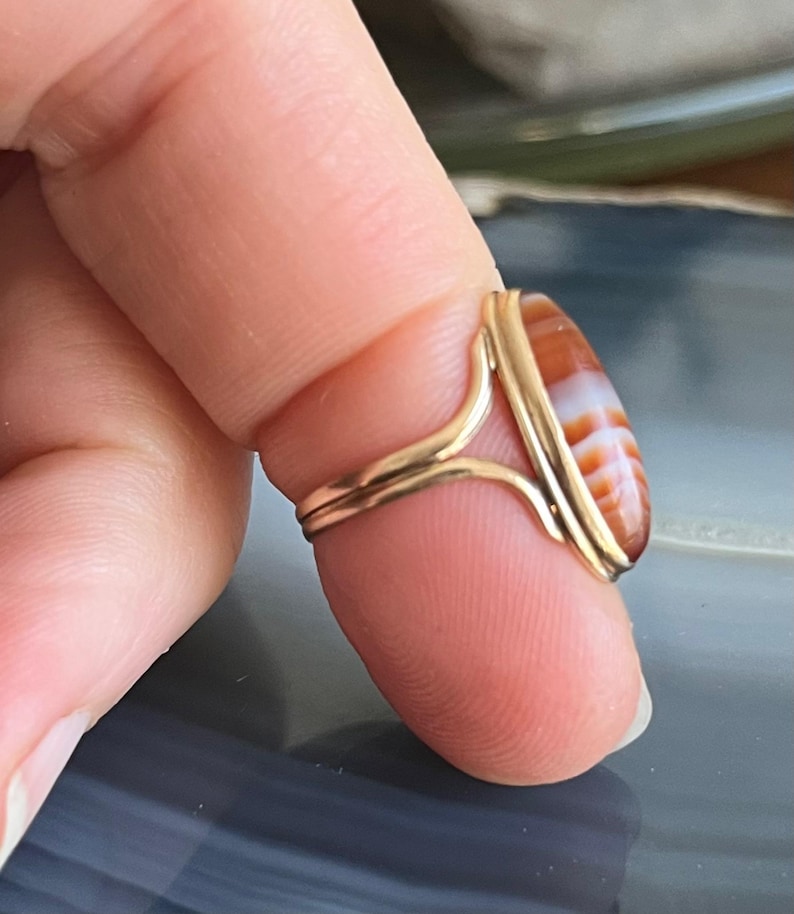 以下が含まれることがあります： 赤と白の瑪瑙の石がベゼルセッティングでセットされた金のリング。リングは指にはめられています。