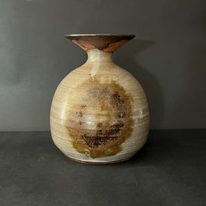 May include: A round ceramic vase with a wide, flared rim and a narrow neck. The body features a light beige base glaze with a prominent, mottled dark brown and olive green circular patch on the front, showing a smooth, glossy finish.