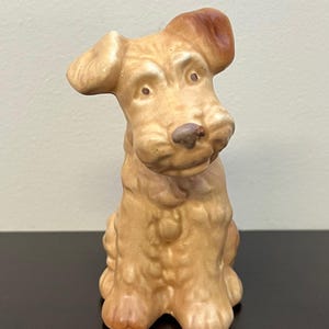 May include: A ceramic dog ornament in a seated pose, with a tilted head and an attentive expression. The dog has a light brown coat with darker brown accents on the ears and nose. The ornament is on a dark surface.
