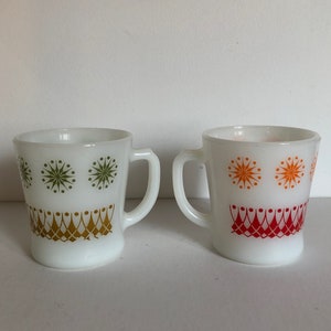 May include: Two white ceramic mugs with geometric designs. One mug has green and gold designs, while the other has orange and red designs.