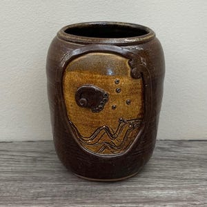 May include: A brown ceramic vase with a decorative design. The vase has a dark brown exterior and a lighter brown interior. The design features a stylized image of a sea creature with waves and bubbles.