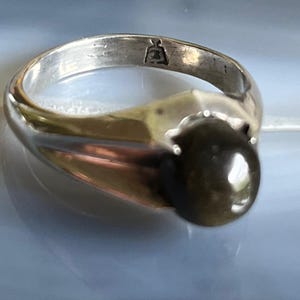 May include: A silver ring with a large, dark, oval-shaped stone. The ring has a simple band and a decorative setting that holds the stone. The ring is on a light blue surface.