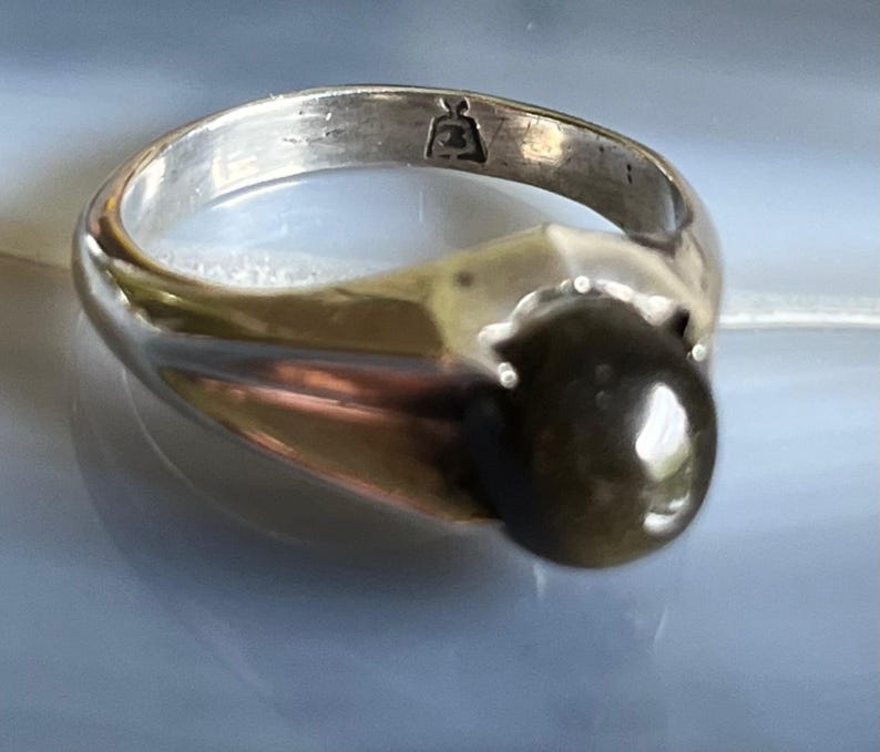 May include: A silver ring with a large, dark, oval-shaped stone. The ring has a simple band and a decorative setting that holds the stone. The ring is on a light blue surface.