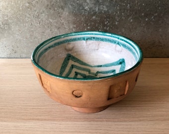 May include: A handmade ceramic bowl with a light brown exterior and a white interior. The rim is accented with a teal stripe, and the interior features teal geometric designs. The bowl has a footed base and decorative cutouts.