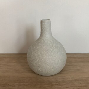 May include: A white ceramic vase with a textured surface. The vase has a round body and a narrow neck.