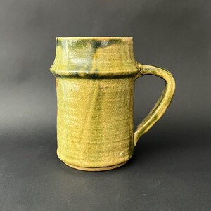 May include: A handmade ceramic mug with a cylindrical body and a curved handle. The mug is glazed in shades of green and yellow, with a textured surface. The mug is approximately 13 cm tall.