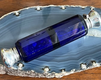 May include: A cobalt blue glass bottle with silver stoppers on both ends. The bottle is sitting on a gray and white agate slice.