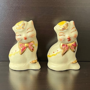 Puede incluir: Dos figuras de gato de cerámica color crema, con los ojos cerrados, pajaritas rojas y detalles dorados. Las figuras de gato son probablemente saleros y pimenteros. Cada gato tiene una cabeza amarilla y dorada.