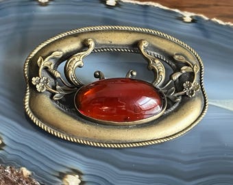 Op de afbeelding: Een vintage broche met een rode cabochon-steen gezet in een messing frame met sierlijke bloemmotieven.