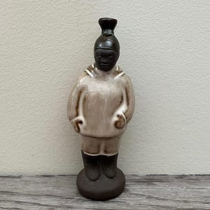 May include: A ceramic figurine of a person in a hooded jacket and shorts. The figurine has a dark brown head and boots, with a light brown body. The figurine is standing on a dark brown circular base.