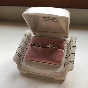 K&ouml;nnte beinhalten: Eine wei&szlig;e Ringbox in Form eines Stuhls mit zwei goldenen Ringen im Inneren. Die Box tr&auml;gt den Text "MEAGHER'S 133 S. Jackson St. Jackson, Mich." auf dem inneren Deckel.