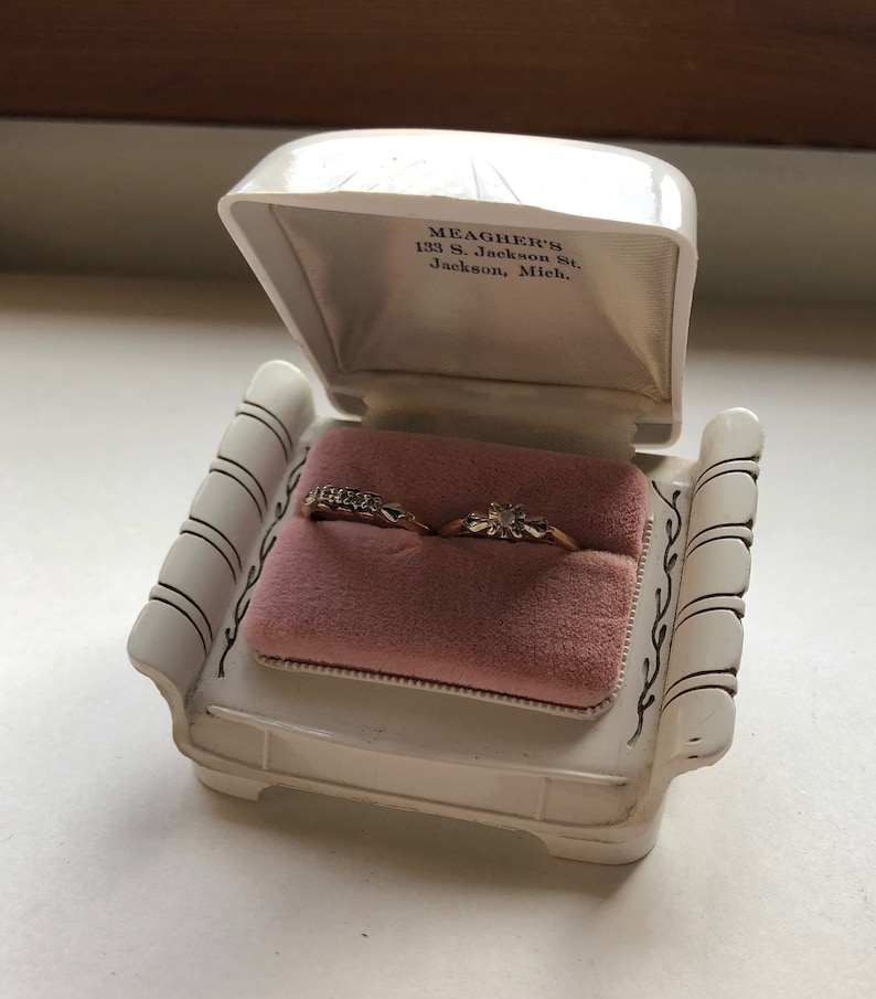 K&ouml;nnte beinhalten: Eine wei&szlig;e Ringbox in Form eines Stuhls mit zwei goldenen Ringen im Inneren. Die Box tr&auml;gt den Text "MEAGHER'S 133 S. Jackson St. Jackson, Mich." auf dem inneren Deckel.