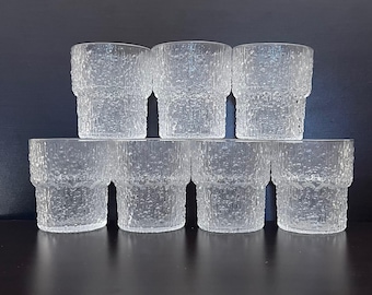 May include: Set of seven clear, textured drinking glasses. Each glass has a cylindrical shape with a textured, bark-like pattern. The glasses are stacked in two rows, with the top row of three glasses and the bottom row of four glasses.