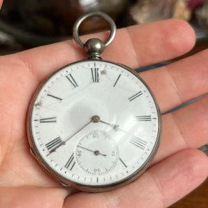 Könnte beinhalten: Antike silberne Taschenuhr mit weißem Zifferblatt und schwarzen römischen Ziffern. Die Uhr hat ein kleineres Hilfszifferblatt und eine Schlaufe oben für eine Kette. Das Zifferblatt ist gerissen. Ein klassischer Zeitmesser.