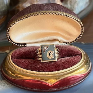Peut inclure: Une bague en or avec une face rectangulaire vert foncé ornée d'un diamant et de la lettre "G". La bague est présentée dans un écrin à bijoux vintage, doublé de velours bordeaux et bordé d'or.
