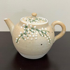 Puede incluir: Una tetera de cerámica beige con tapa, boquilla y asa. La tetera está decorada con diseños florales blancos y verdes. La tetera está sobre una superficie marrón oscuro.