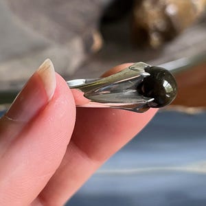 May include: A silver ring featuring a dark, rounded gemstone. The ring's setting has a unique, stylized design that cradles the stone. The gemstone appears to be polished and reflects light, creating a subtle shine. The ring is held by a person's fingers.