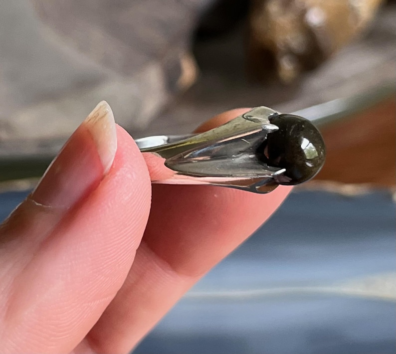 May include: A silver ring featuring a dark, rounded gemstone. The ring's setting has a unique, stylized design that cradles the stone. The gemstone appears to be polished and reflects light, creating a subtle shine. The ring is held by a person's fingers.