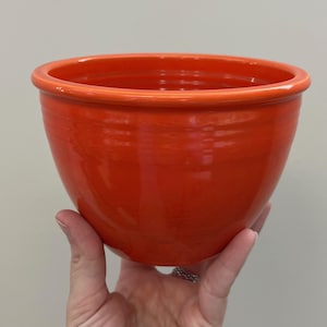 Könnte beinhalten: Eine leuchtend orangefarbene Keramikschüssel mit glänzendem Finish. Die Schüssel hat eine abgerundete Form mit einem leicht ausgestellten Rand. Die Schüssel wird von einer Hand gehalten, die ihre Größe und ihr Design zeigt. Diese Schüssel eignet sich zum Servieren oder als dekoratives Stück.