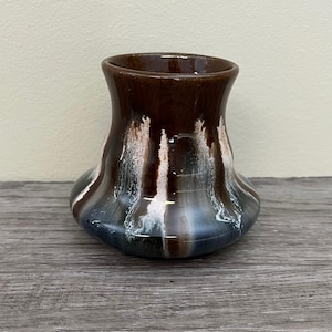 Peut inclure: Petit vase en céramique fait à la main avec un glaçage unique. Le vase présente un dessus brun foncé qui se transforme en stries blanches et bleues, créant un effet de goutte. Le vase a une base arrondie et une ouverture légèrement évasée. Le vase est posé sur une surface en bois.