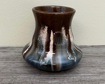 Pode incluir: Um pequeno vaso de cerâmica artesanal com uma esmaltação única. O vaso apresenta um topo marrom escuro que faz a transição para listras brancas e azuis, criando um efeito de gotejamento. O vaso tem uma base arredondada e uma abertura ligeiramente alargada. O vaso é exibido em uma superfície de madeira.