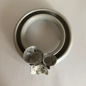 May include: A white ceramic wall hanging with a ring shape and three white ceramic flowers attached to the bottom.