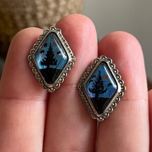 May include: A pair of silver earrings with a blue and black butterfly wing design featuring a silhouette of a tree. The earrings are shaped like a diamond.