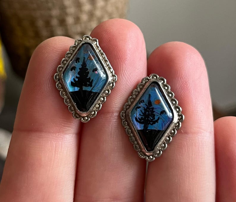 May include: A pair of silver earrings with a blue and black butterfly wing design featuring a silhouette of a tree. The earrings are shaped like a diamond.