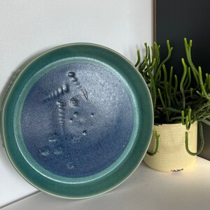 Peut inclure: Une assiette en céramique bleue et verte avec un motif texturé. L'assiette a un bord surélevé et un centre profond.