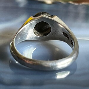 May include: A silver ring with a dark, possibly black, stone set in a triangular bezel. The ring's band is smooth and rounded, with markings visible on the inside. The ring is displayed against a blurred, light blue and white background.