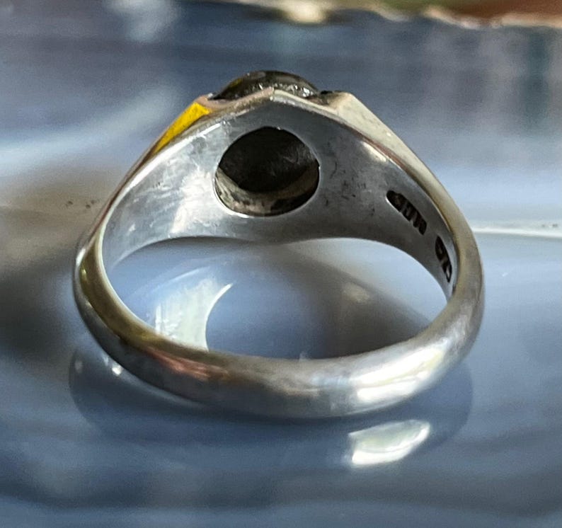 May include: A silver ring with a dark, possibly black, stone set in a triangular bezel. The ring's band is smooth and rounded, with markings visible on the inside. The ring is displayed against a blurred, light blue and white background.