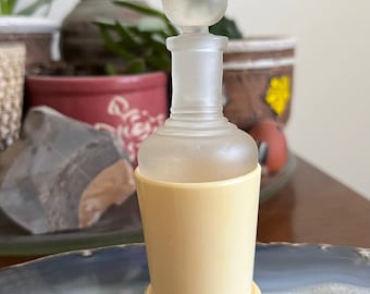 May include: A clear glass bottle with a frosted finish and a bulbous stopper. The bottle is sitting on a pale yellow base and is resting on a blue and gray agate slice.