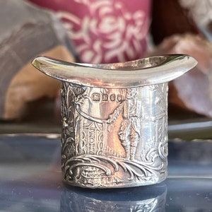 May include: A silver top hat shaped trinket with an engraved design of a man in a top hat and a building. The top hat has the inscription "TH" and a hallmark.