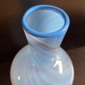 May include: A decorative glass vase with a light blue body and a flared neck. The vase features a swirl pattern of blue, red, and white stripes. The rim of the vase is a solid light blue. The vase has a rounded base.