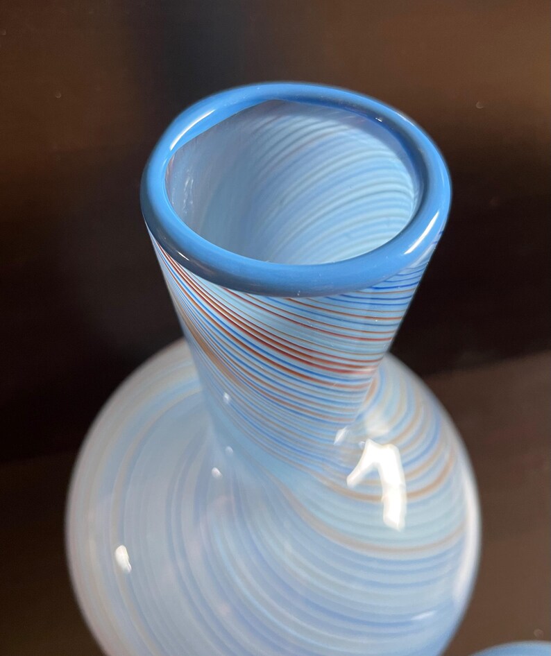 May include: A decorative glass vase with a light blue body and a flared neck. The vase features a swirl pattern of blue, red, and white stripes. The rim of the vase is a solid light blue. The vase has a rounded base.