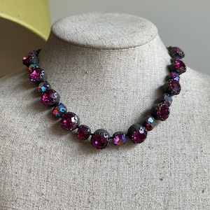 May include: A striking necklace with large, round, dark purple crystals and iridescent highlights. The necklace is strung on a black cord and displayed on a mannequin. The crystals are a deep magenta colour, making it a bold piece of jewellery.