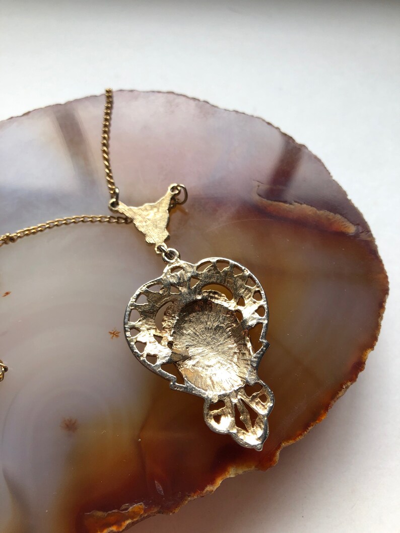 May include: A gold pendant necklace with a detailed filigree design. The pendant is shaped like a teardrop and has a circular cutout in the center. The necklace is on a gold chain.