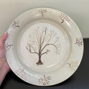 May include: A beige ceramic plate with a hand-painted tree design. The tree is brown with delicate branches, centered on the plate. Smaller tree branch designs are spaced around the rim. The plate has a speckled texture.