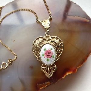 May include: A gold necklace with a delicate chain and a pendant featuring a white enamel oval with a pink rose and green leaves.