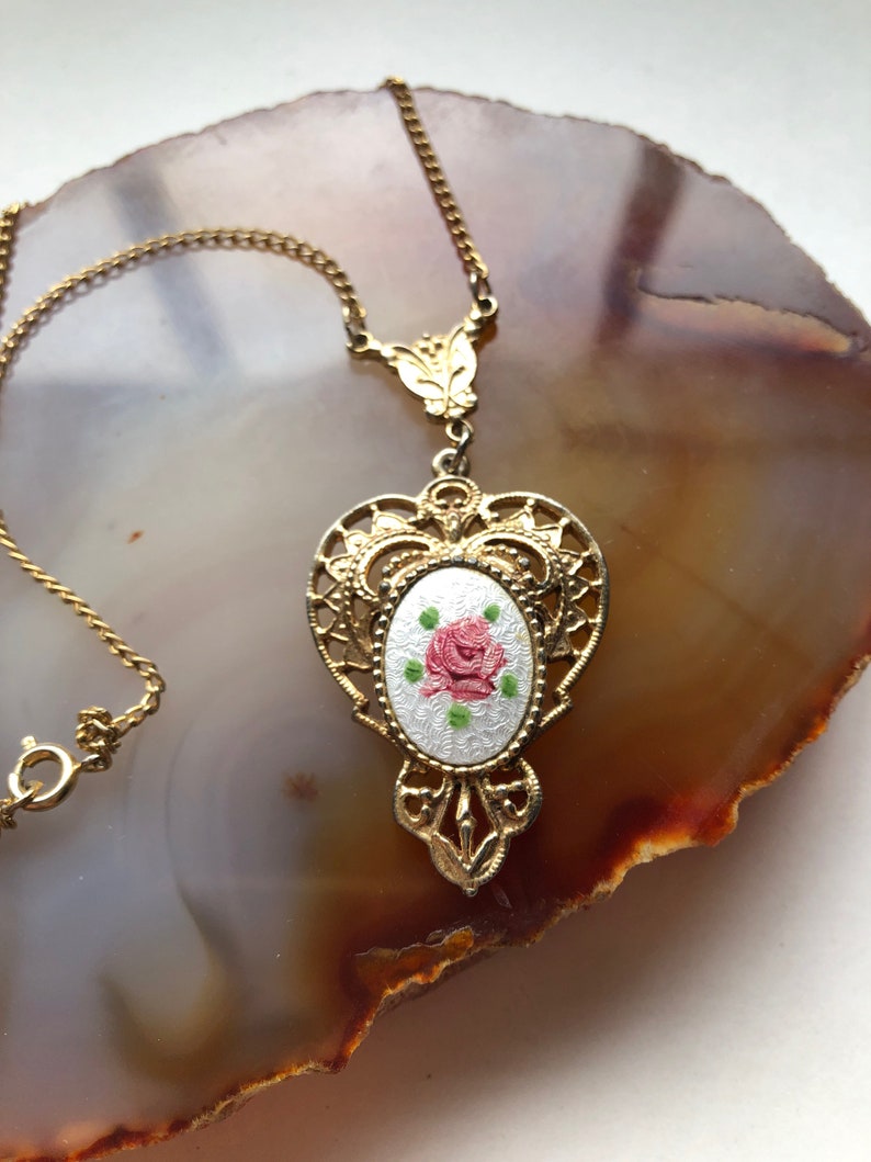 May include: A gold necklace with a delicate chain and a pendant featuring a white enamel oval with a pink rose and green leaves.