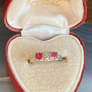 Peut inclure: Une bague vintage avec deux pierres précieuses rondes rouge vif et de petites pierres d'accent, serties dans un anneau argenté. La bague est présentée dans un écrin rouge en forme de cœur avec des inscriptions dorées.