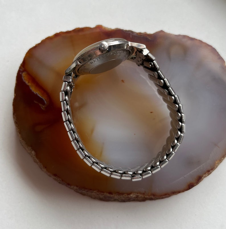 May include: A silver wristwatch with a silver metal band. The watch face is round and has a silver rim. The watch is on a brown and white agate surface.