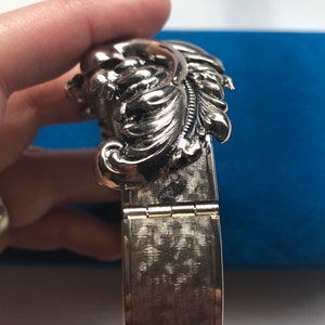 K&ouml;nnte beinhalten: Ein silbernes Armband mit einem dekorativen Blumenmuster. Das Armband ist mit einem Scharnier versehen und hat eine strukturierte Oberfl&auml;che.